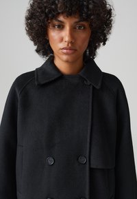 Cappotto nero doppiopetto con colletto, realizzato in un tessuto texturizzato, con bottoni e una vestibilità leggermente rilassata.