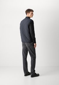 Vestă matlasată de culoare bleumarin cu guler înalt, purtată peste o cămașă neagră, asortată cu blugi gri și pantofi negri, prezentând un design minimalist.