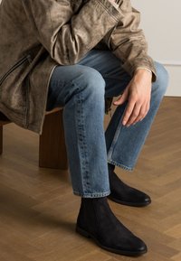 Zwarte suède enkellaarzen met elastische zijpanelen, gedragen met lichtblauwe rechte jeans en een gestructureerde bruine leren jas.