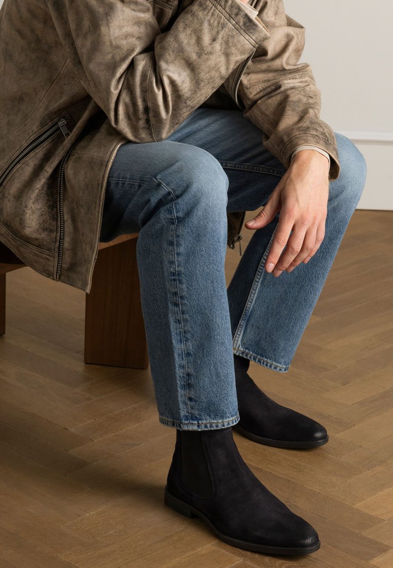 Zwarte suède enkellaarzen met elastische zijpanelen, gedragen met lichtblauwe rechte jeans en een gestructureerde bruine leren jas.