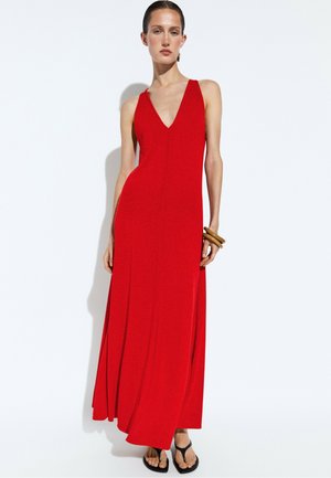 Donna che indossa un vestito lungo rosso senza maniche con scollatura a V, sandali neri e braccialetti di legno, in piedi su sfondo bianco.