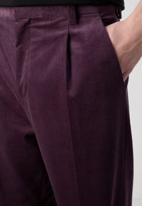 Pantalons en velours côtelé violet avec une texture lisse, comportant des plis avant et des poches. La taille est plate, et le tissu présente un léger côtelé.