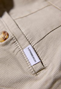 Tissu à carreaux beige avec une surface texturée, présentant un bouton et une étiquette blanche avec le texte "LINDERGH" en noir.