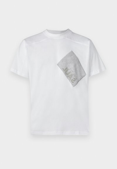 Valkoinen puuvillapaita, jossa on tekstiilinen hopeinen timanttilappu vasemmalla puolella. Lyhyet hihat ja klassinen pyöreä kaula-aukko.