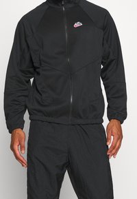 Man in een zwarte Nike zip-up jacket met elastische manchetten en bijpassende zwarte broek, staand tegen een effen lichte achtergrond.