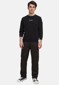 Zwarte sweatshirt met wit logo op de borst, gecombineerd met donkergroene broek en zwart-witte sneakers. Model staat tegen een witte achtergrond.