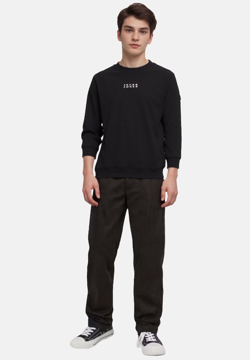Zwarte sweatshirt met wit logo op de borst, gecombineerd met donkergroene broek en zwart-witte sneakers. Model staat tegen een witte achtergrond.