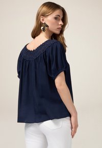 Blusa blu navy con scollatura in pizzo texturizzato, maniche a palloncino corte e vestibilità ampia. Abbinata a pantaloni bianchi.