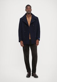 Homem em pé, vestido com um casaco pea navy, suéter castanho com fecho, calças castanhas escuras e botas de couro castanho escuras, contra um fundo simples.