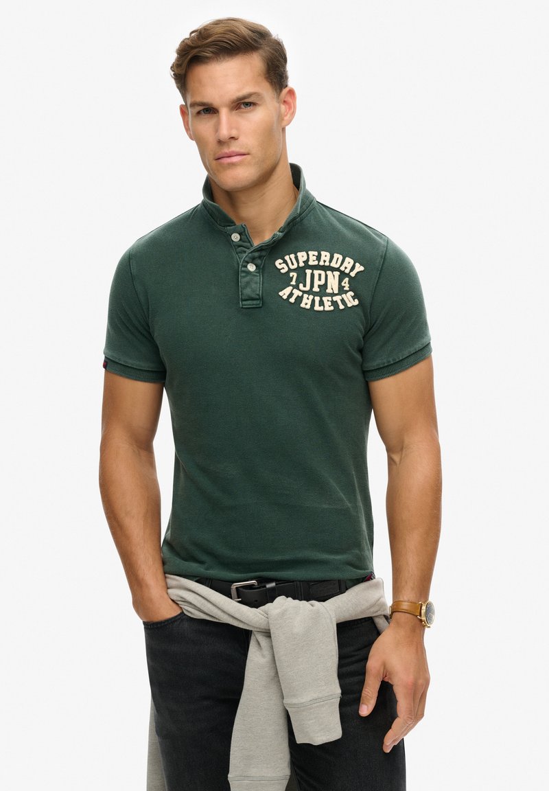 Donkergroen polo shirt met een geribbelde textuur, voorzien van een drie-knopen sluiting en vet witte letters. Grijze sweatshirt rond de taille geknoopt.