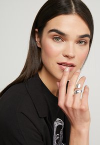 Silver metal ring with a smooth, shiny surface; two bands intertwine. Model has long dark hair, wearing a black shirt with a graphic design.