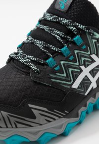 Baskets de sport noires avec une tige texturée, ornées d'accents zigzag turquoise et blancs, de lacets ronds et d'une semelle extérieure multicolore pour une meilleure adhérence.