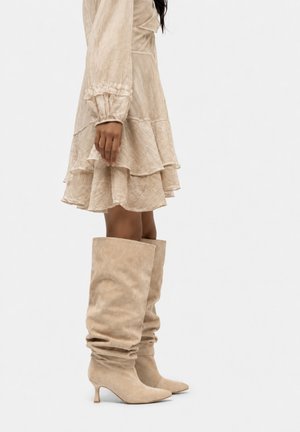 Vrouw draagt een beige getextureerde lange mouwen jurk met ruches en bijpassende kniehoge slordige laarzen met lage hakken.