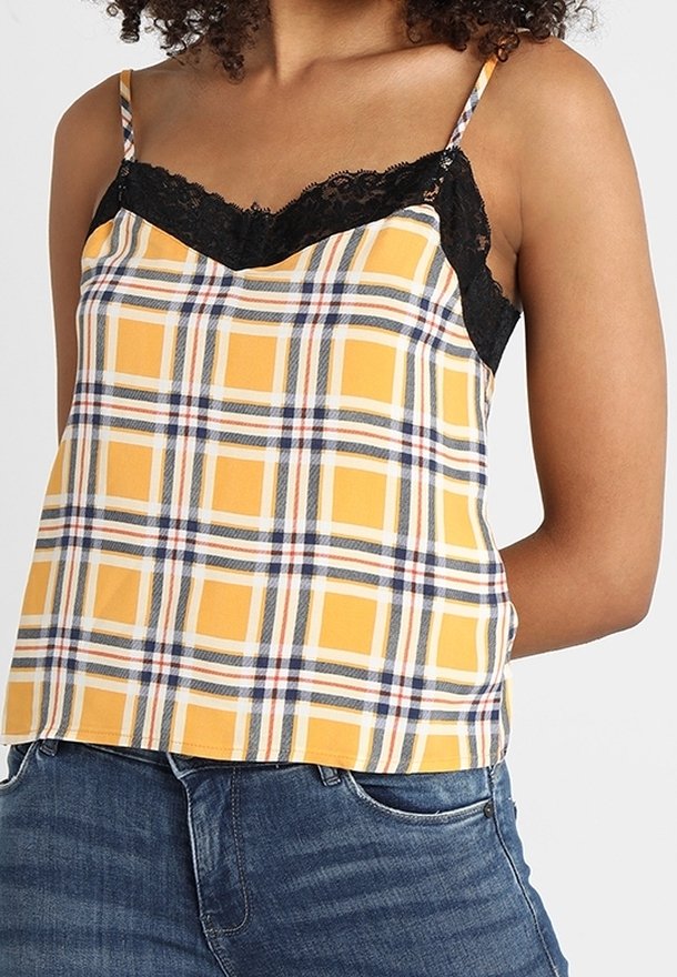 Femme portant un caraco à carreaux jaunes avec bordure en dentelle noire et un jean bleu, mains derrière le dos, sur un fond uni clair.