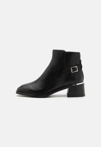 Bottine en cuir noir avec un bout pointu, un accent argenté sur le talon et un détail de sangle réglable sur le côté.
