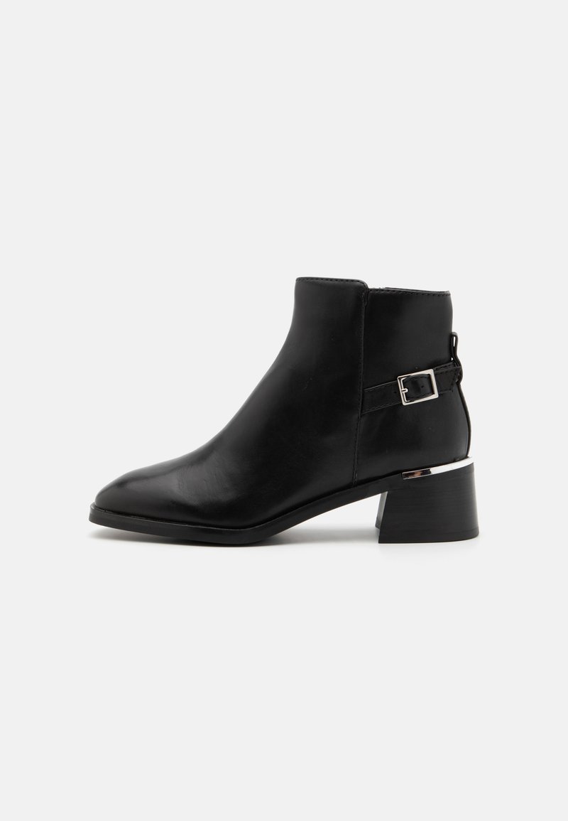 Bottine en cuir noir avec un bout pointu, un accent argenté sur le talon et un détail de sangle réglable sur le côté.
