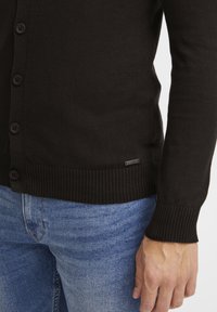 Mężczyzna w czarnej zapinanej kardiganie i niebieskich dżinsach, z widoczną dłonią po boku.