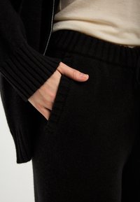 Černý pletený svetr a kalhoty, s žebrovanými manžetami a pasem, s rukou položenou v kapse kalhot, viditelná měkká textura.