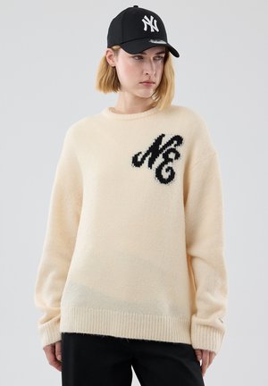 Nainen, jolla on päällään kerma neule, jossa mustat kirjaimet "N" ja "G", sekä musta New York Yankees -lippalakki, seisoo yksiväristä taustaa vasten.