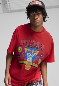 Tricou roșu din bumbac, cu un design grafic ce include un coș de baschet, "PUMA BASKETBALL" și accente colorate. Poartă cu pantaloni scurți cu modele.