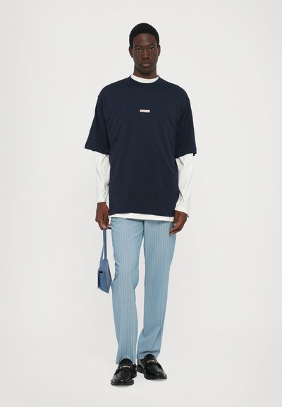 Marinblå oversized t-shirt lagd över en vit långärmad skjorta, parat med ljusblå randiga byxor och svarta loafers, hållande en blå väska.