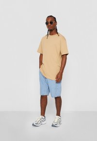 Un tricou oversized de culoare bej, pantaloni scurți din denim de un albastru deschis, adidași argintii și ochelari de soare rotunzi negri. Design simplu, fără modele sau logo-uri.