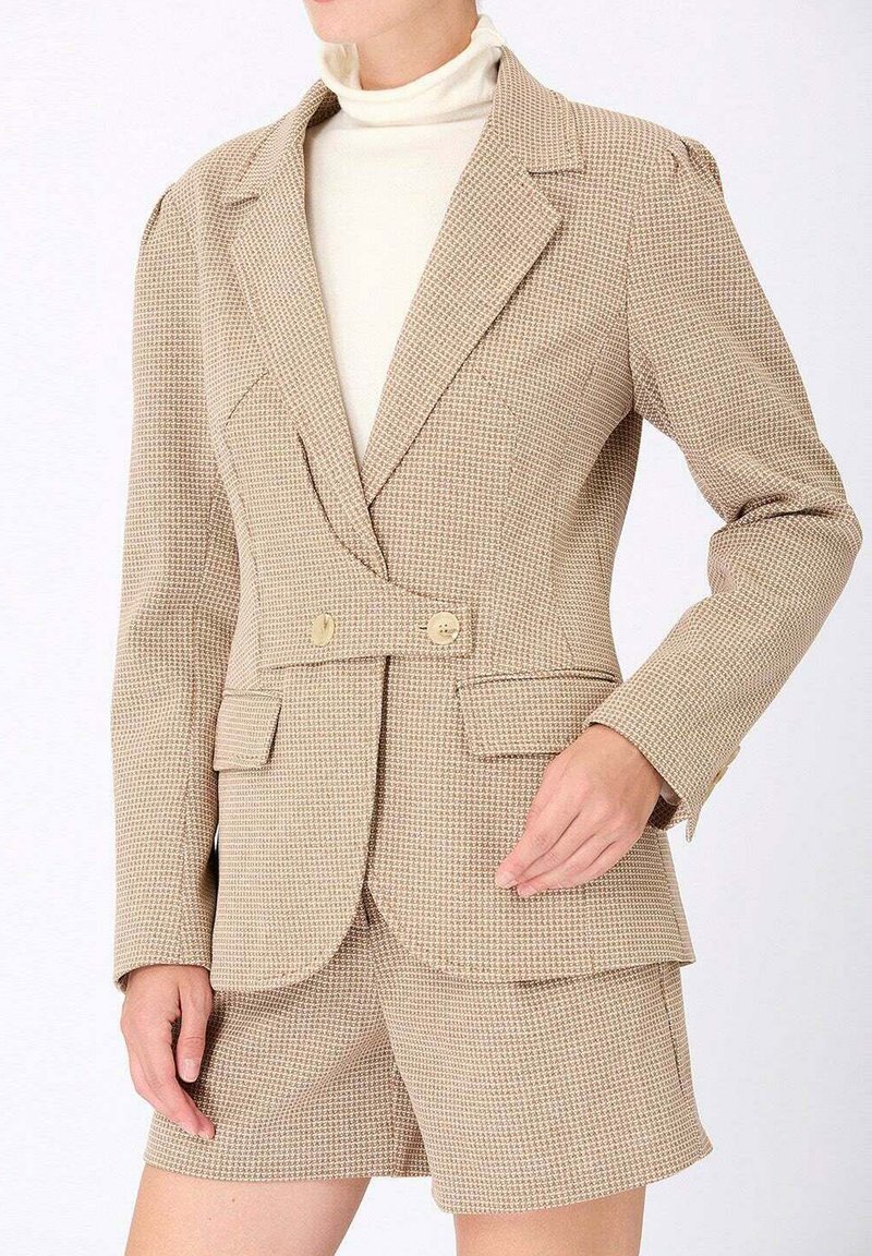 Blazer beige en pied-de-poule avec double boutonnage, revers aigus et tissu texturé, associé à un short taillé assorti.