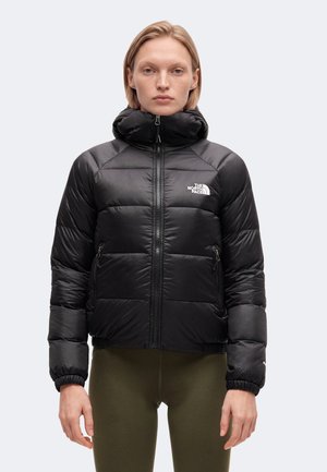 Άτομο που φοράει μαύρο ενισχυμένο μπουφάν North Face με ψηλό γιακά και φερμουάρ μπροστά, συνδυασμένο με παντελόνι ελιάς, στέκεται μπροστά σε απλό φόντο.