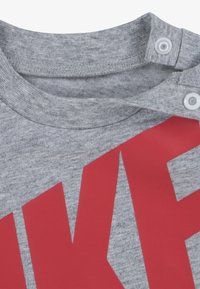 Γκρι βαμβακερό μπλουζάκι με κόκκινο γράφημα "NIKE". Διαθέτει στρογγυλή λαιμόκοψη, δύο κουμπιά στον ώμο και απαλή υφή.