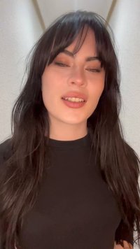 Jeune femme aux longs cheveux foncés avec une frange, portant un haut noir, souriant légèrement devant un mur uni de couleur claire.