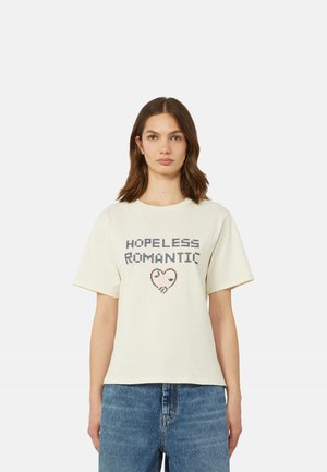 Donna con capelli castani alla lunghezza delle spalle indossa una maglietta bianca con la scritta "HOPELESS ROMANTIC" e un cuore pixellato, abbinata a jeans blu.