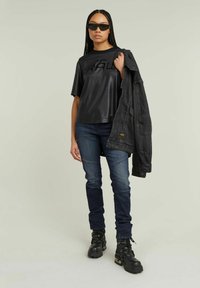 Must jupp musta "RAW" logo, tume teksad, mustad lahingukingi ja must denim-jakk, mis on õlal. Päikeseprillid täiendavad välimust.