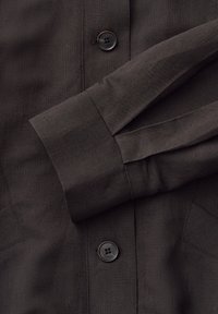 Donkerbruine blouse met lange mouwen, knoopmanchetten en zwarte knopen aan de voorkant, gemaakt van gestructureerde geweven stof.