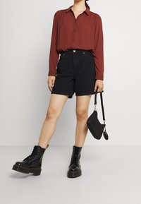 Shorts en jean noirs courts associés à un chemisier rouille boutonné. La personne tient un petit sac  à main noir. Des bottes de combat noires brillantes complètent la tenue.