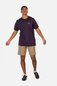 Camiseta de algodón púrpura, de manga corta, con cuello redondo. Pantalones cortos beige, hasta la rodilla, sin bolsillos. Zapatillas de correr negras con detalles en blanco. Fondo liso.