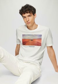 Witte katoenen t-shirt met een rechthoekige afbeelding van een auto op een weg tegen een woestijnlandschap. Model gecombineerd met lichtcrèmekleurige broeken.