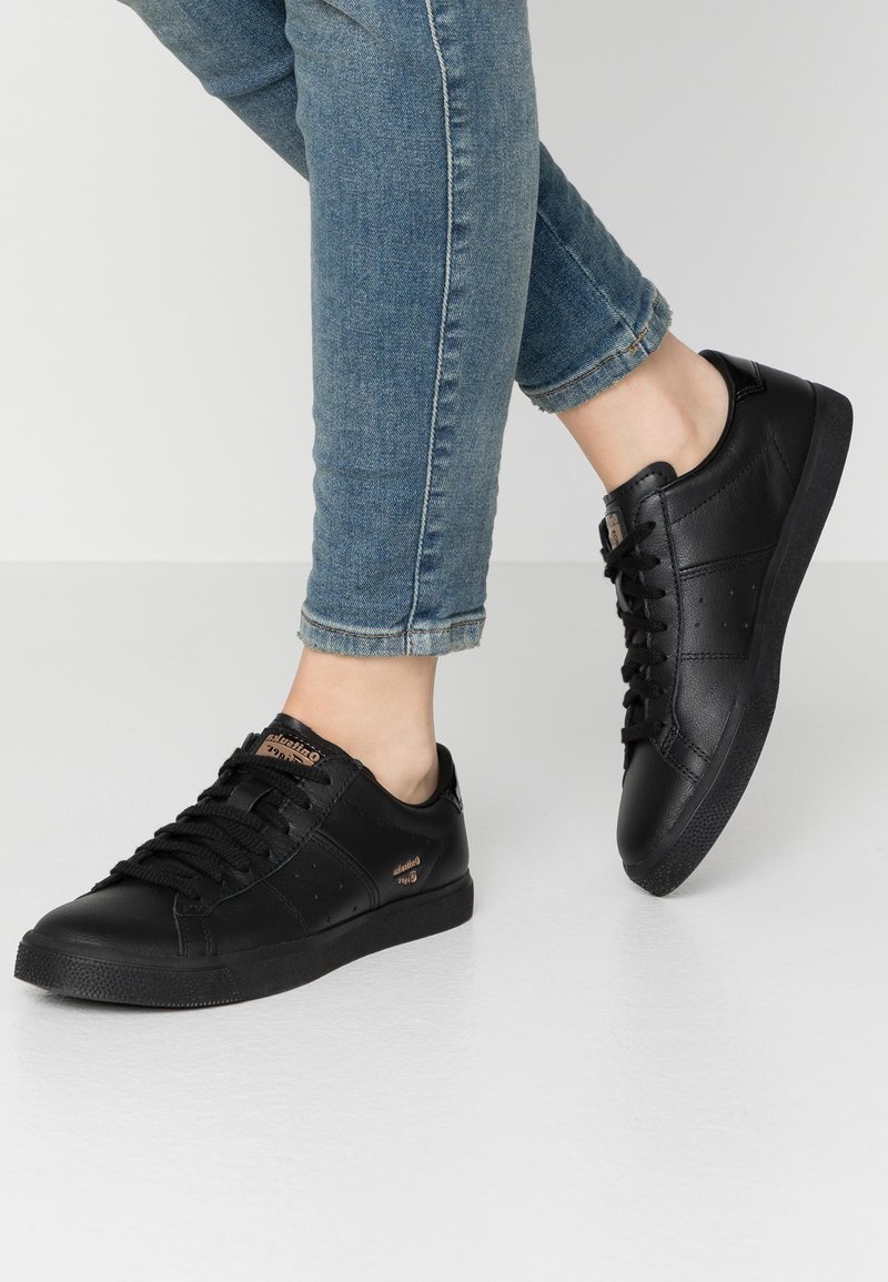 Black leather sneakers with flat laces, textured toe cap, and perforated side detailing. Worn with light blue cuffed jeans.