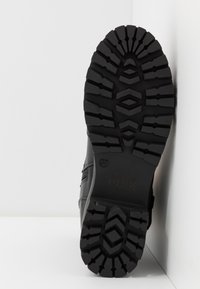 Semelle de botte noire avec un motif de bande de roulement profond et robuste, présentée sur un fond blanc avec angles.