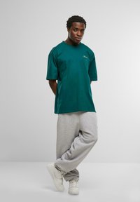 Tricou din bumbac verde-albastru, cu o croială lejeră, asociat cu pantaloni de trening gri și adidași albi. Prezintă un mic logo auriu pe tricou.