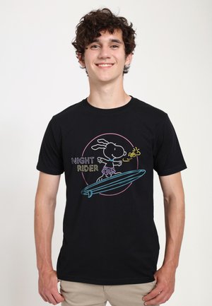 Ung mand smiler, iført en sort T-shirt med et neon-stil Snoopy surfing design og ordene "Night Rider."