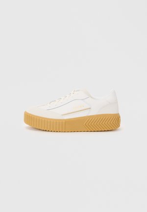 Hvit sneaker med en teksturert tan gummisåle, med et glattere overmateriale og subtil merkedekorasjon i gull på siden.