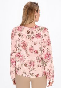 Blusa floreale in tessuto rosa trasparente con maniche lunghe, spalle arricciate e retro con grandi stampe di fiori rosa e verdi.