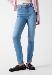Jean skinny en denim bleu clair, à taille haute, texture lisse et design classique à cinq poches. Associé à des mocassins noirs.