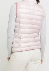 Hellrosa gesteppte Weste mit horizontaler Steppung, hohem Kragen und ärmellosem Design. Wird über einem grauen Langarmshirt getragen, mit minimalen Akzenten.