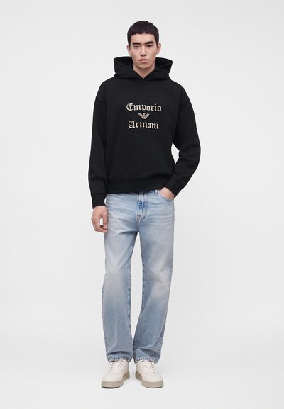 Νέος άνδρας που φοράει μαύρο φούτερ Emporio Armani, ανοιχτό μπλε τζιν και λευκά αθλητικά παπούτσια, στέκεται με το ένα χέρι στην τσέπη.