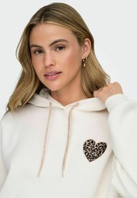 Hvit hettegenser med et leopardmønstret hjertepatch på fronten. Den har snøring med en teksturert, metallisk finish.