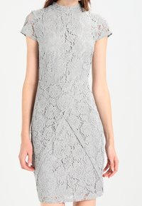 Harmaa pitsimekko lyhyillä hihoilla, korkealla kaula-aukolla, kukkakuviolla, tiiviillä siluetilla ja vinolla viimeistelyllä hameessa.