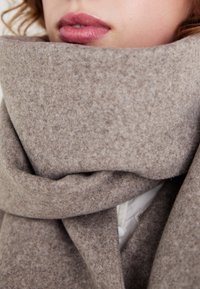 Beige ullskjerf med en myk tekstur, med et tykt, drapert design. Nærbilde som viser jevne fibre og naturlige fargevariasjoner.