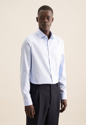 Chemise bleu clair en coton, avec un col évasé et un devant à boutons. Manches longues avec des poignets boutonnés, associée à un pantalon foncé.