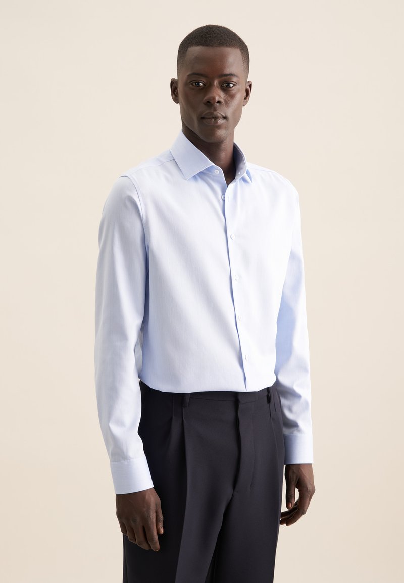 Camicia azzurra in cotone, con colletto alla francese e chiusura a bottoni. Maniche lunghe con polsini a botone, abbinata a pantaloni scuri.